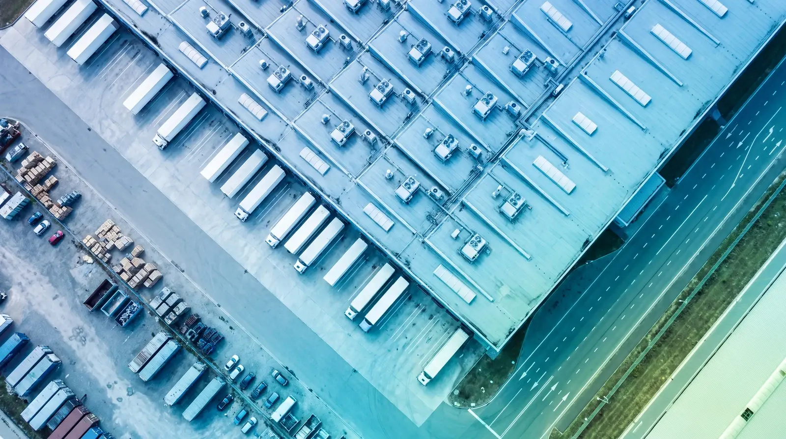 Overhead view of a manufacturing plant roof with rooftop units and skylights and a row of trailers at the docks, in a blue-teal grade
